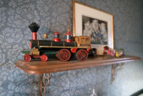 A worn wooden toy train displayed on a shelf. The engine is labeled "Hercules" and is pulling three colorful cars.
