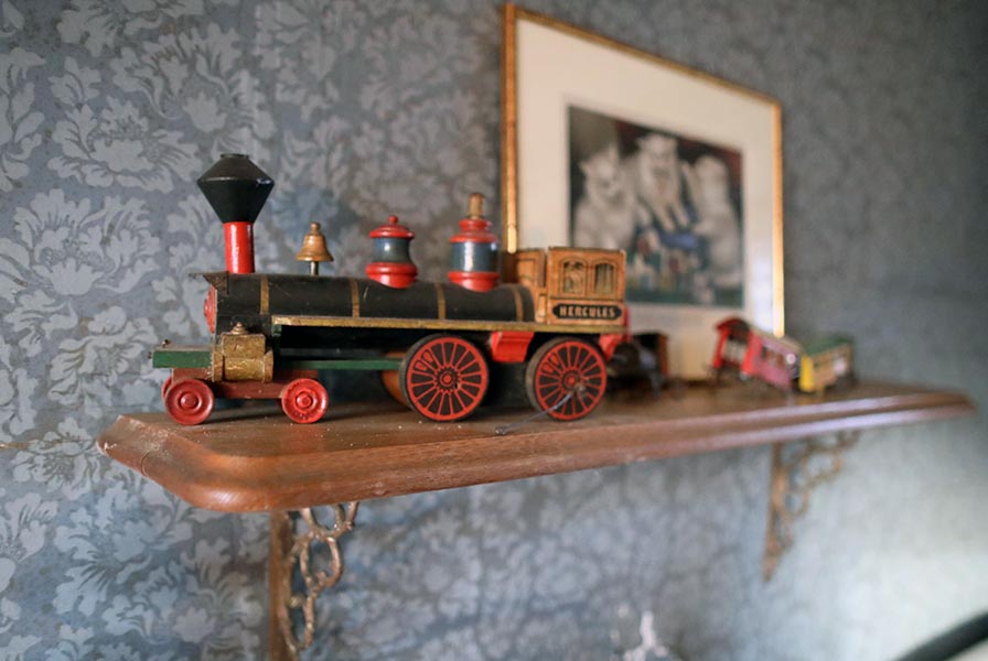 A worn wooden toy train displayed on a shelf. The engine is labeled "Hercules" and is pulling three colorful cars.