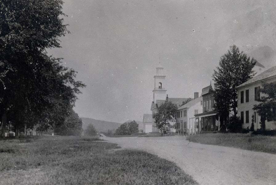 A row of buildings along a dirt road. The two-storey post office has a pillared porch and decorative timber framing.