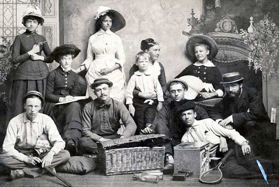 10 young adults and child pose casually before a painted backdrop. In front are a tennis racket, bottles and a wicker chest.