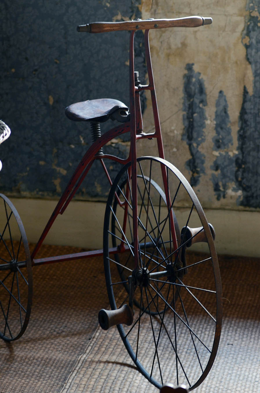 A velocipede (an early tricycle with a large front wheel). It has a red frame, wooden handlebars and pedals, and narrow wheels.