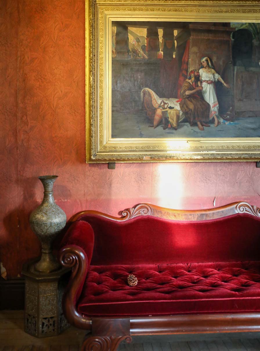 A gilt-framed oil painting of Abram and Sara hangs above a red velvet sofa. To the left, is a brass vase on a taboret