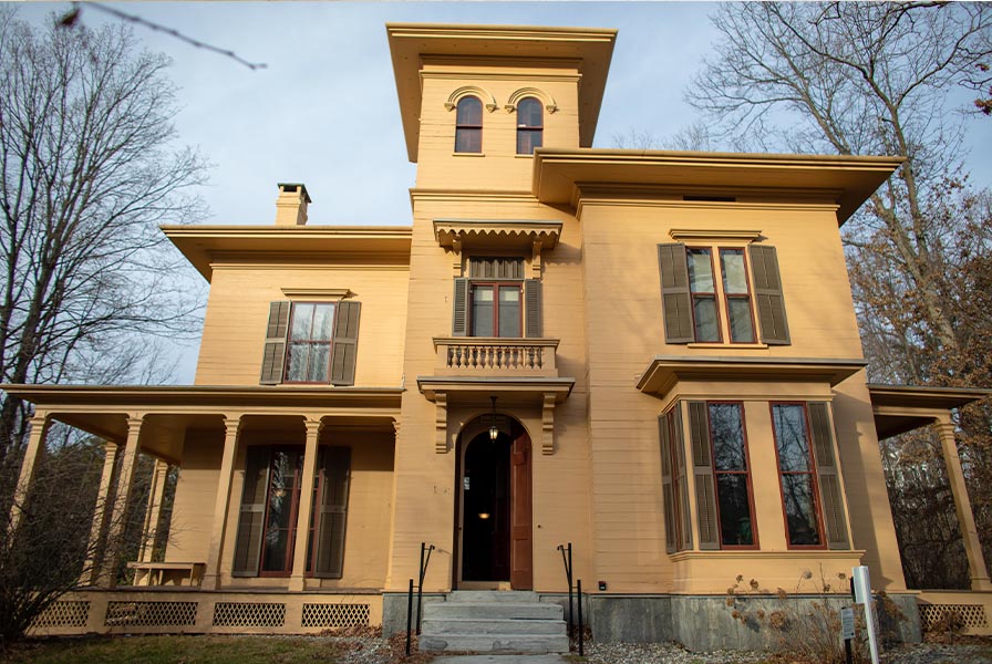 Ochre-colored Italianate-style house with overhanging eaves and a square cupola. The many windows have green shutters.