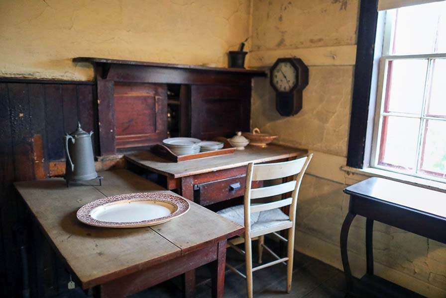 In the corner of the kitchen are work tables laid with ceramic serving dishes. A wallclock is hung next to a sunlit window