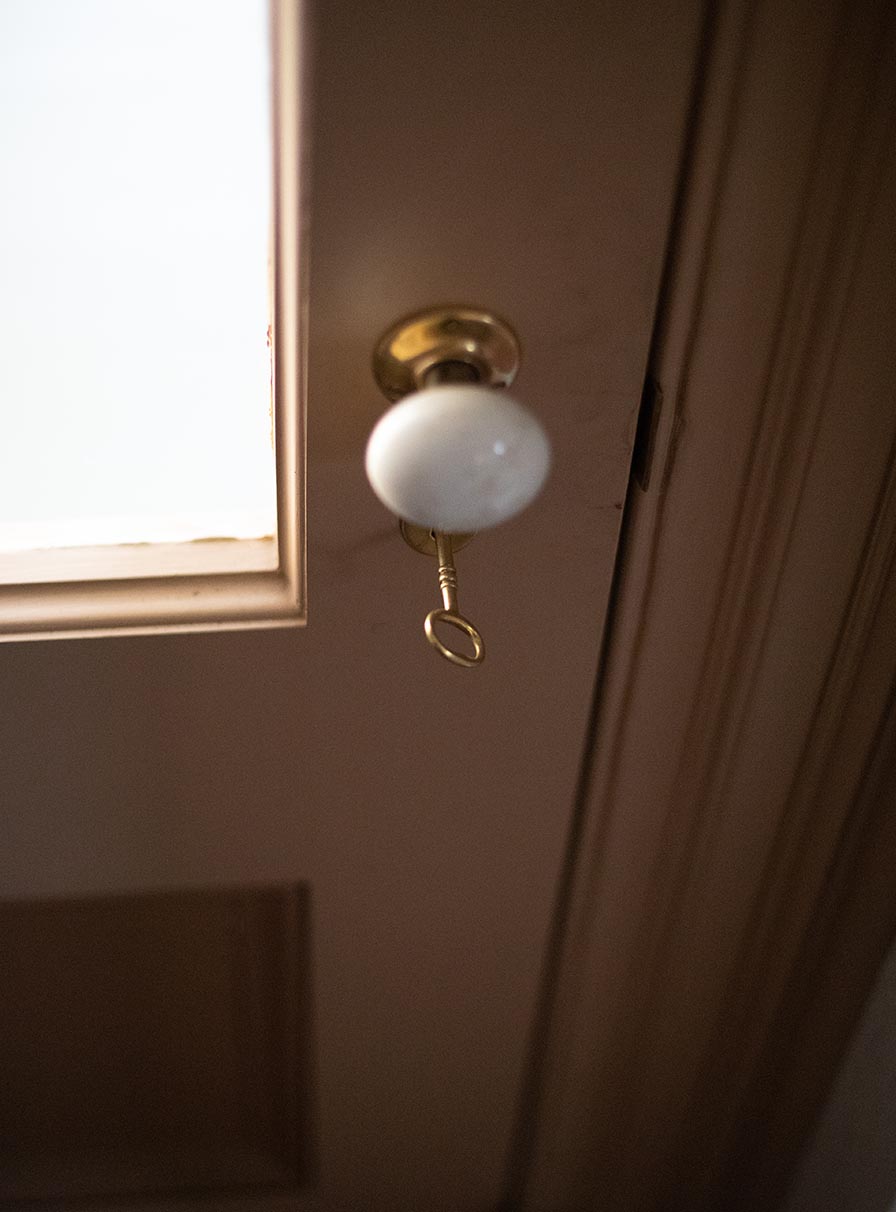 A bright brass key turned in the lock of a tan door with white ceramic doorknob and clear glass panel.