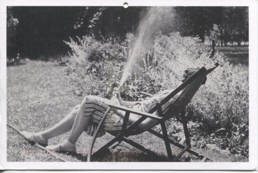 Black and white postcard depicting a photograph of Eudora Welty sitting outside by a sprinkler and a garden