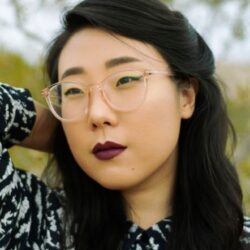 Photo of Franny Choi, looking away from the camera with one arm behind her head. She has long black hair that is pulled back behind her ears, dark red lipstick and clear glasses.