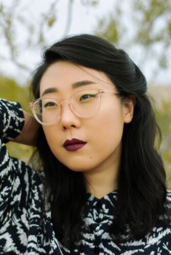 Photo of Franny Choi, looking away from the camera with one arm behind her head. She has long black hair that is pulled back behind her ears, dark red lipstick and clear glasses.