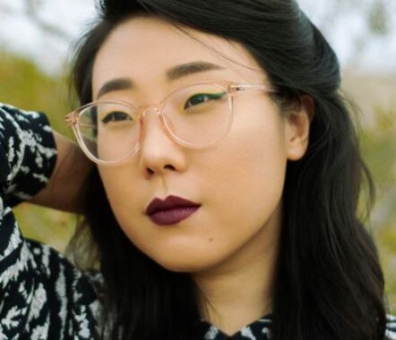 Photo of Franny Choi, looking away from the camera with one arm behind her head. She has long black hair that is pulled back behind her ears, dark red lipstick and clear glasses.