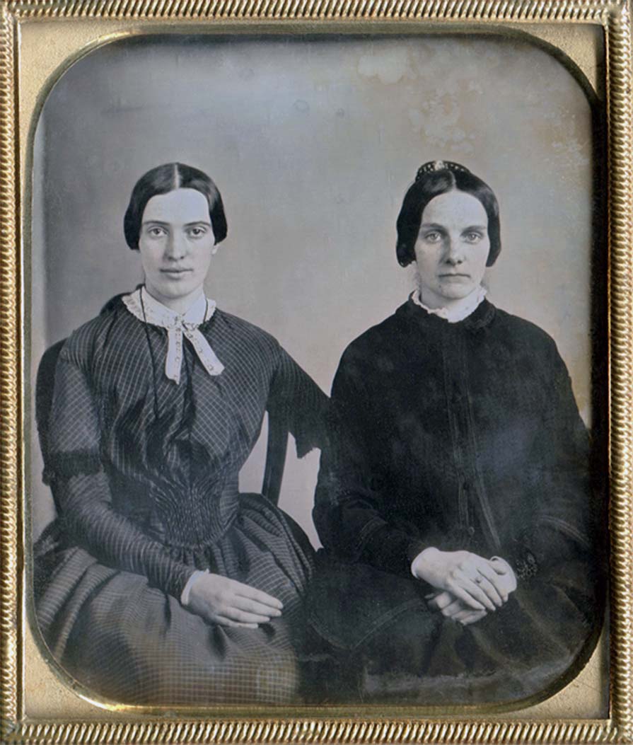 Two seated women in dresses. The one to the left (possibly Dickinson) is smiling slightly with her arm behind the other