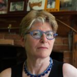 a woman with short blonde hair, blue glasses, and blue necklace stands in front a fireplace