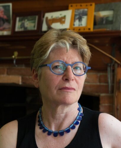 a mature woman with short blonde hair, blue glasses, and blue necklace stands in front a fireplace