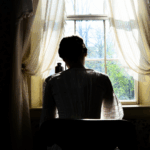 a model dressed as Dickinson with her back to the camera sitting at her writing desk