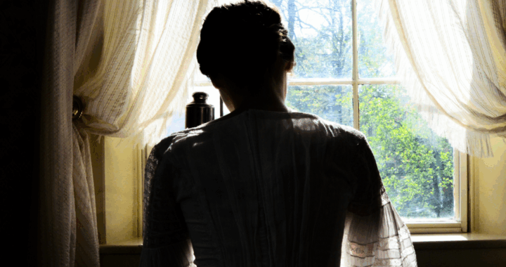 a model dressed as Dickinson with her back to the camera sitting at her writing desk