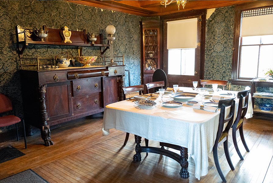 Well-appointed dining room with corner cabinets, claw-footed table and sideboard. The wallpaper is a blue brocade pattern.